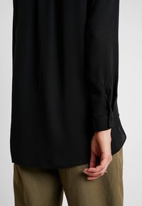 Blusa nera a maniche lunghe con una vestibilità comoda, caratterizzata da polsini con bottone e spacchi laterali, mostrata su uno sfondo chiaro.