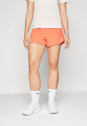 Personne portant des chaussures de sport blanches, des chaussettes Nike blanches, un short de course Nike corail et une chemise blanche, debout sur un fond clair.