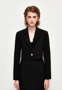 Zkrácený černý blazer s límečkem, jedním zlatým knoflíkem, hladkou texturou, spárovaný s přiléhavými černými šaty.