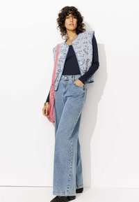 Gilet à motifs bleus sur un haut à manches longues navy, associé à un jean large bleu clair et un sac à main rose, avec des chaussures à clous noires.