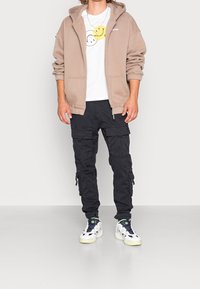 Sudadera beige con cremallera, camiseta blanca con gráfico de cara sonriente, pantalones cargo negros con bolsillos laterales y zapatillas blancas con detalles verdes.