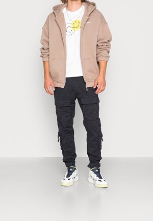 Beżowa bluza na zamek, biały t-shirt z grafiką uśmiechniętej buźki, czarne spodnie cargo z bocznymi kieszeniami oraz białe trampki z zielonymi akcentami.