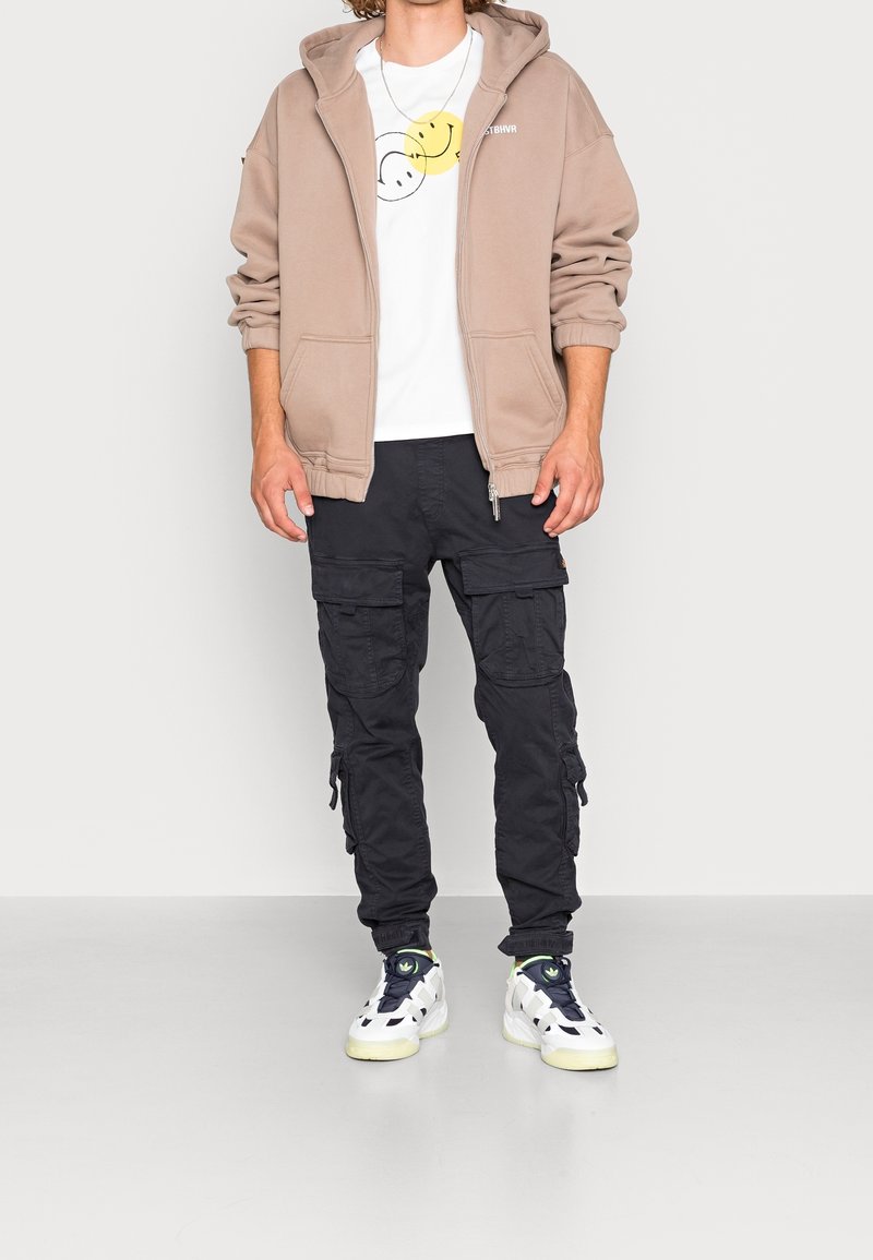 Sudadera beige con cremallera, camiseta blanca con gráfico de cara sonriente, pantalones cargo negros con bolsillos laterales y zapatillas blancas con detalles verdes.