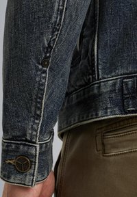 Veste en denim bleu foncé avec une texture délavée, présentant un poignet à boutons et des coutures contrastantes beiges, associée à un pantalon vert olive.