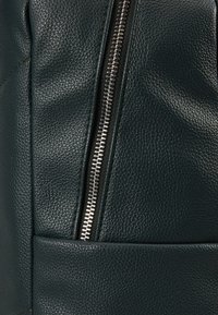 Gros plan sur une veste en cuir texturé vert foncé avec une fermeture éclair en métal argenté et une couture en cuir horizontale près du bas.