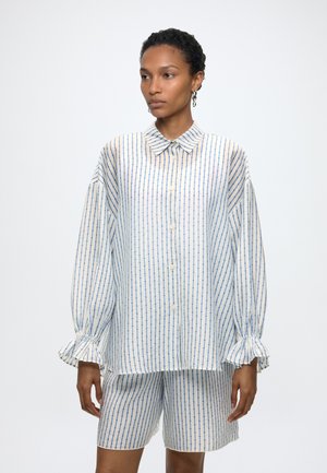 Frau mit kurzen Haaren, die ein locker sitzendes weißes Hemd und passende Shorts mit vertikalen blauen Streifen trägt, steht vor einem einfarbigen Hintergrund.