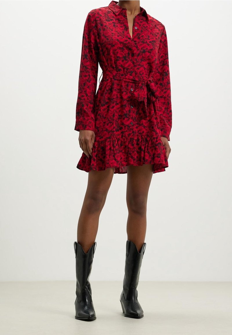 Femme portant une robe-chemise longue à manches longues rouge à fleurs avec une ceinture à la taille et des bottes de cowboy noires à mi-mollet, debout sur un fond uni.