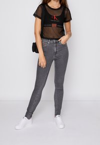 Černé průsvitné síťované tričko s červeným logem Calvin Klein, kombinované s přiléhavými šedými džíny a bílými teniskami; model stojí na jednotném pozadí.