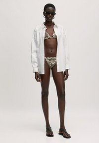 Witte oversized shirt, gedragen over een bruin en beige zebra-gestreepte bikini, gecombineerd met zwarte flip-flops en een zonnebril.