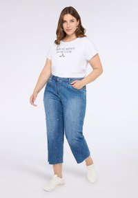 Camiseta blanca con texto negro, combinada con jeans de mezclilla azules que presentan adornos multicolor. El modelo lleva zapatillas blancas.