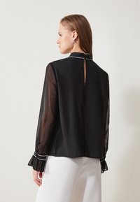 Blusa nera trasparente con maniche lunghe, dettagli in bianco e apertura a forma di chiave sulla parte posteriore. I polsini svasati aggiungono una sottile texture. Indossata sopra pantaloni bianchi.
