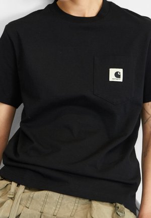 Person iført en sort Carhartt t-shirt med brystlomme og beige cargo-bukser, synlig fra halsen til taljen mod en lys baggrund.