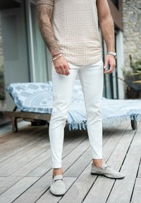 Chemise à motifs beige clair, pantalon blanc ajusté, mocassins en suède gris. Le décor présente une terrasse en bois et des meubles aux accents bleus en arrière-plan.
