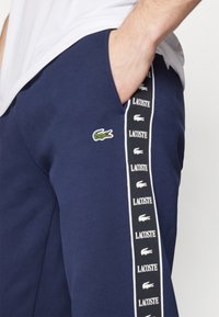 Granatowe spodnie dresowe wykonane z miękkiej tkaniny, z bocznymi kieszeniami, małym zielonym logo Lacoste oraz białą taśmą z paskami i logo marki wzdłuż boków.