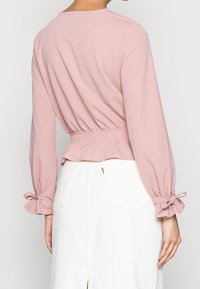 Blusa rosa a maniche lunghe con vita arricciata e maniche legate, realizzata in morbido tessuto testurizzato. Indossata con una gonna bianca al ginocchio.