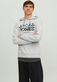 Sudadera con capucha gris que presenta un gran estampado de logotipo negro y de camuflaje en el frente, combinada con pantalones negros. Diseño simple y ajuste cómodo.