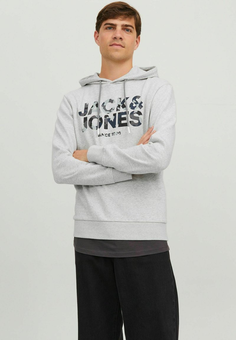 Sudadera con capucha gris que presenta un gran estampado de logotipo negro y de camuflaje en el frente, combinada con pantalones negros. Diseño simple y ajuste cómodo.