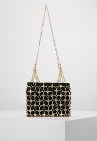 Bolso rectangular negro con revestimiento de red de cadena dorada y doble asa de cadena dorada, expuesto sobre una superficie blanca.