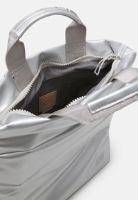 Stříbrná metalická taška s dvojitými uchy, zipovým uzávěrem a texturovaným povrchem. Uvnitř se nachází zipová kapsa a logo štítek.