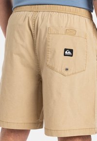 Shorts bej cu talie elastică, buzunar la spate și un logo de marcă negru. Materialul pare neted, având o croială casual și lejeră.