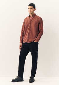 Chemise à boutons couleur rouille avec une poche poitrine, associée à un pantalon noir et des chaussures foncées sur un fond neutre.