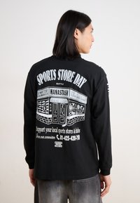 Black long-sleeve t-shirt featuring white graphic text and illustration on the back, promoting "Sports Store Day" with storefront details.