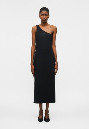 Kobieta ubrana w czarną, midi sukienkę na jedno ramię i czarne szpilki w szpic, stojąca na tle jednolitego jasnego tła.