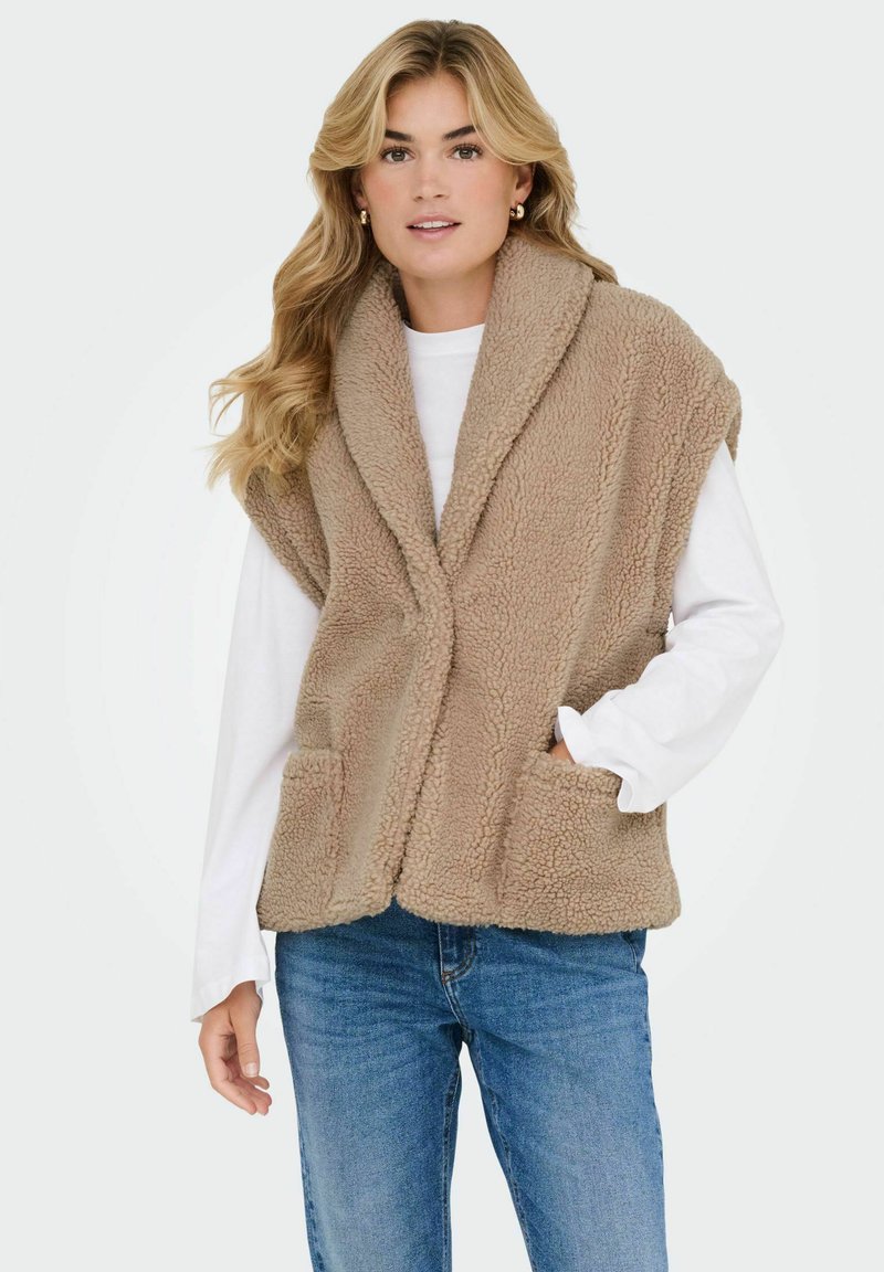 Bežna, puhasta fleece brezrokavnik s šalom, odprtim sprednjim delom in sprednjimi žepi, nošena čez belo majico z dolgimi rokavi, v kombinaciji z modrimi kavbojkami.