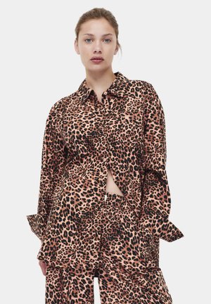 Junge Frau mit zurückgebundenem Haar trägt ein locker sitzendes Leopardenmuster-Shirt und passende Hose vor einem schlichten hellen Hintergrund.