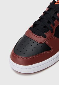 Lærsneaker med en bordeauxfarget overdel, svart perforert tåhette og hvit gummisåle. Har svarte snørebånd og oransje logodetalj.