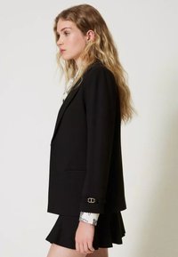Zwarte getailleerde blazer met een notch lapel, enkele knoopsluiting en decoratieve manchetbewerking over een gestreept wit shirt. Gecombineerd met een zwarte rok.