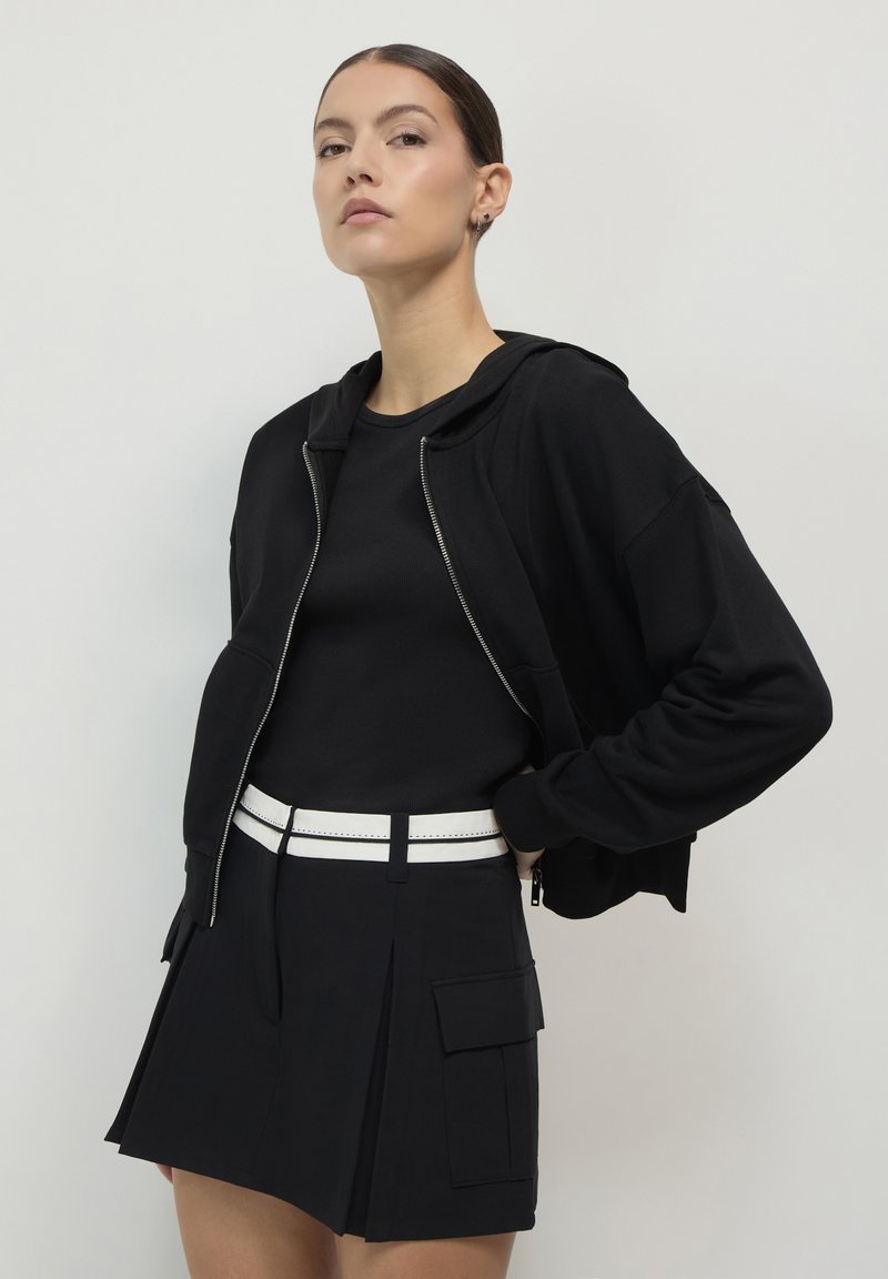Jeune femme avec les cheveux tirés en arrière portant un sweat à capuche noir zippé, un haut noir et une jupe noire avec une ceinture blanche, posant sur un fond uni.