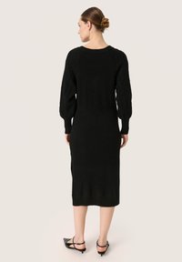 Rochie neagră tricotată cu mâneci lungi, cu nervuri și o siluetă ajustată. Textura prezintă un model cu cabluri pe umeri. Design până la gleznă.