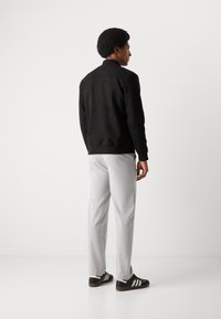 Black suede jacket with a ribbed collar, paired with light gray trousers and black sneakers featuring three white stripes.