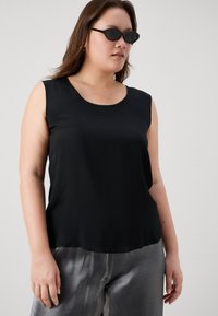 Svart ermeløs topp i glatt stoff med rund hals, kombinert med blanke sølvbukser. Modellen har på seg mørke, kantete solbriller.