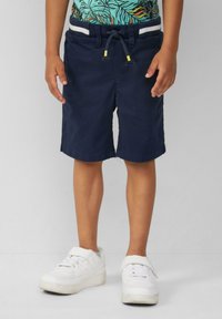 Pantaloni shorts blu navy con cintura elastica, cordino e righe bianche. Abbinati a una camicia con stampa tropicale e sneaker bianche.