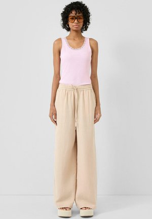 Kobieta o kręconych włosach, nosząca pomarańczowe okulary przeciwsłoneczne, różowy top bez rękawów, beżowe spodnie z szerokimi nogawkami oraz sandały na platformie, stojąca na jednolitym tle.