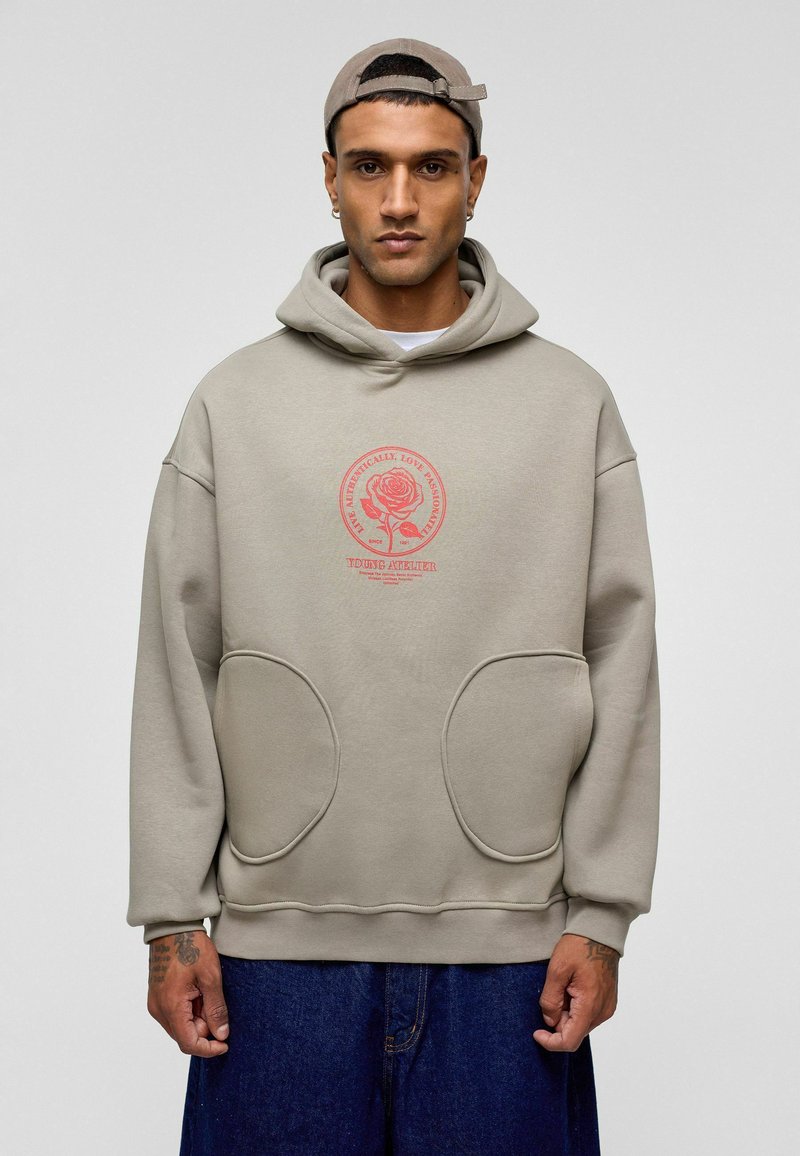 Beige oversized hoodie met een grote grafische rozenprint in het roze. Voorzien van een kap met trekkoord, twee oversized voorkboeftzingen en geribbelde manchetten.