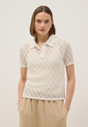 Donna che indossa una polo lavorata a maglia bianca a maniche corte con motivo a diamante e gonna beige, in piedi con le mani nelle tasche su sfondo uniforme.