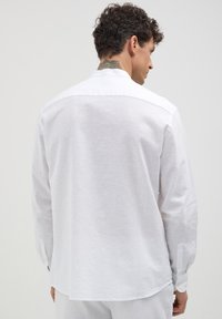 Mann mit dunklen, lockigen Haaren und Nackentattoo, der von hinten vor einem schlichten Hintergrund in einem langen weißen Hemd mit Knöpfen zu sehen ist.