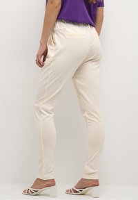 Pantalon couleur crème, fuselé, avec une taille élastique, deux poches arrière et une texture lisse et douce. Associé à des sandales blanches à talons hauts.