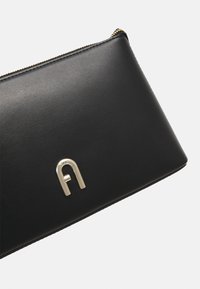 Clutch rettangolare in pelle nera con cerniera dorata e emblema logo metallico sul davanti su sfondo bianco.