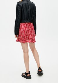 Giacca corta in pelle nera sopra una minigonna rossa a fiori con orlo arricciato, abbinata a sandali neri con zeppa, vista da dietro.