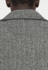 Juodos ir baltos spalvos striukė su žuvies kaulų raštu, turinti struktūruotą apykaklę ir tekstūruotą audinį, matoma iš nugaros.