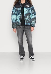 Gewatteerd pufferjack met een blauw-zwart tie-dye patroon, voorzien van een hoge kraag, ritssluiting en een logo op de linkerborst. Gecombineerd met grijze jeans en sneakers.