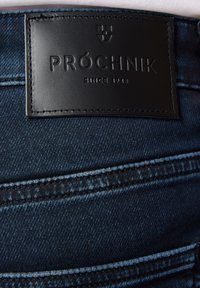 Ciemnoniebieskie dżinsy z czarną skórzaną naszywką z wytłoczonym napisem "PRÓCHNIK" oraz "SINCE 1948". Widoczne szwy i detale tekstury.