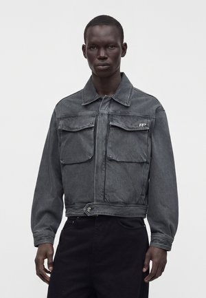 Homme portant une veste en jean noire délavée avec deux poches poitrine et un petit logo blanc sur la poche gauche, associée à un pantalon noir.