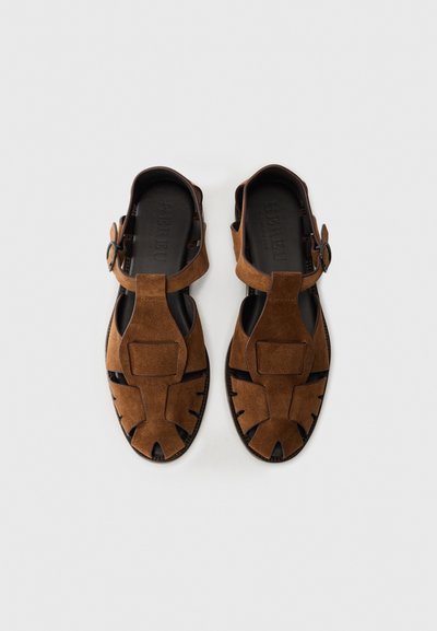 Brown suede sandals with a closed toe, cut-out design, adjustable strap, and rubber sole. Features wide front and textured accents.