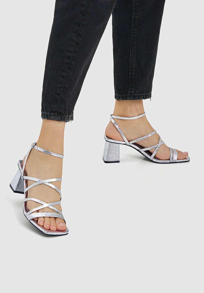Pieds portant des sandales argentées à talons blocs et brides, associées à un pantalon noir court, sur un fond uni.