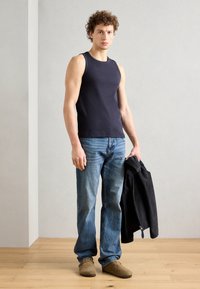 Top fără mâneci de culoare bleumarin, blugi de denim, și pantofi slip-on din suedă maro, cu o jachetă neagră purtată pe braț, stând pe un podea din lemn.